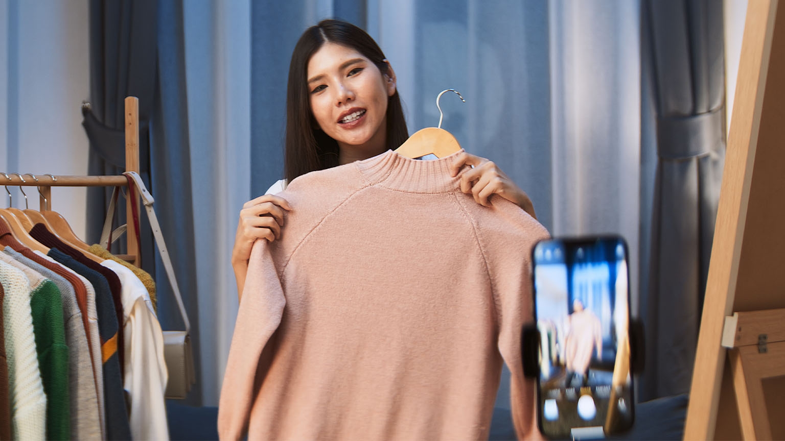 A woman holds a peach colored sweater and records a video on her phone.